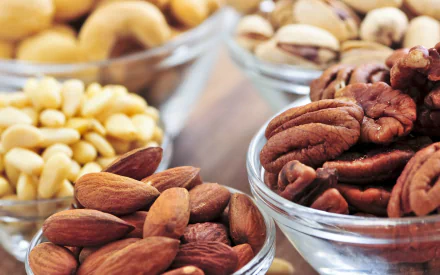 A vibrant HD wallpaper featuring an assortment of nuts in clear bowls, showcasing almonds, pecans, cashews, and pine nuts, set on a wooden surface.