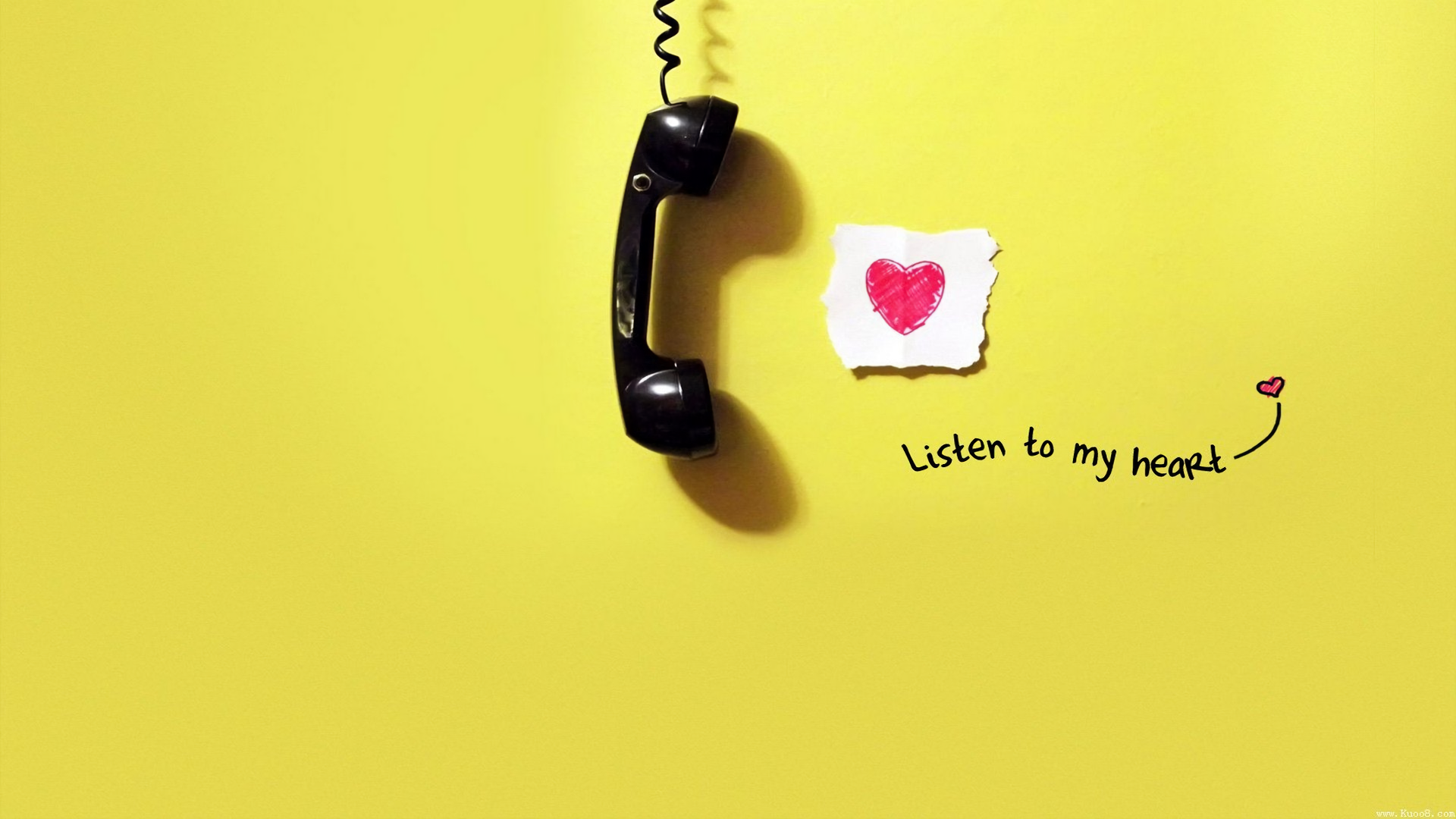 A black telephone hangs against a bright yellow background, next to a note with a heart and the words Listen to my heart, capturing an artistic expression of love.