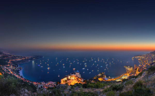 HD desktop wallpaper of Monaco's vibrant coastline and illuminated cityscape against a sunset backdrop, featuring a panoramic ocean view dotted with yachts.