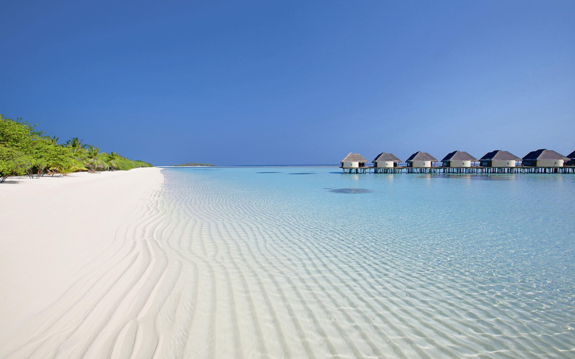 HD PC desktop wallpaper featuring serene beach with clear water, white sand, and a row of huts extending over the ocean under a clear blue sky.