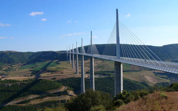 Man-made Millau Viaduct cable-stayed bridge spanning a sunlit green valley beneath a clear blue sky — HD PC desktop wallpaper and background.