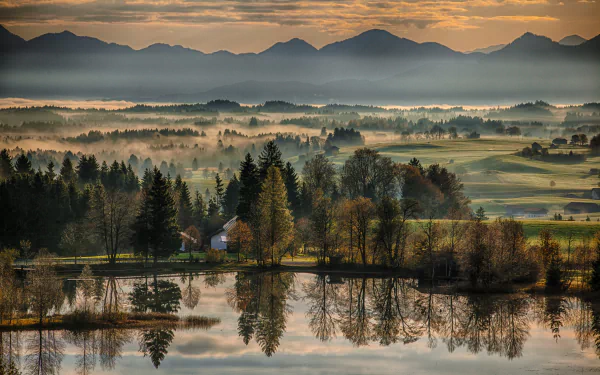 HD desktop wallpaper featuring a misty landscape with mountains in the background and trees reflecting in a tranquil lake at sunrise.