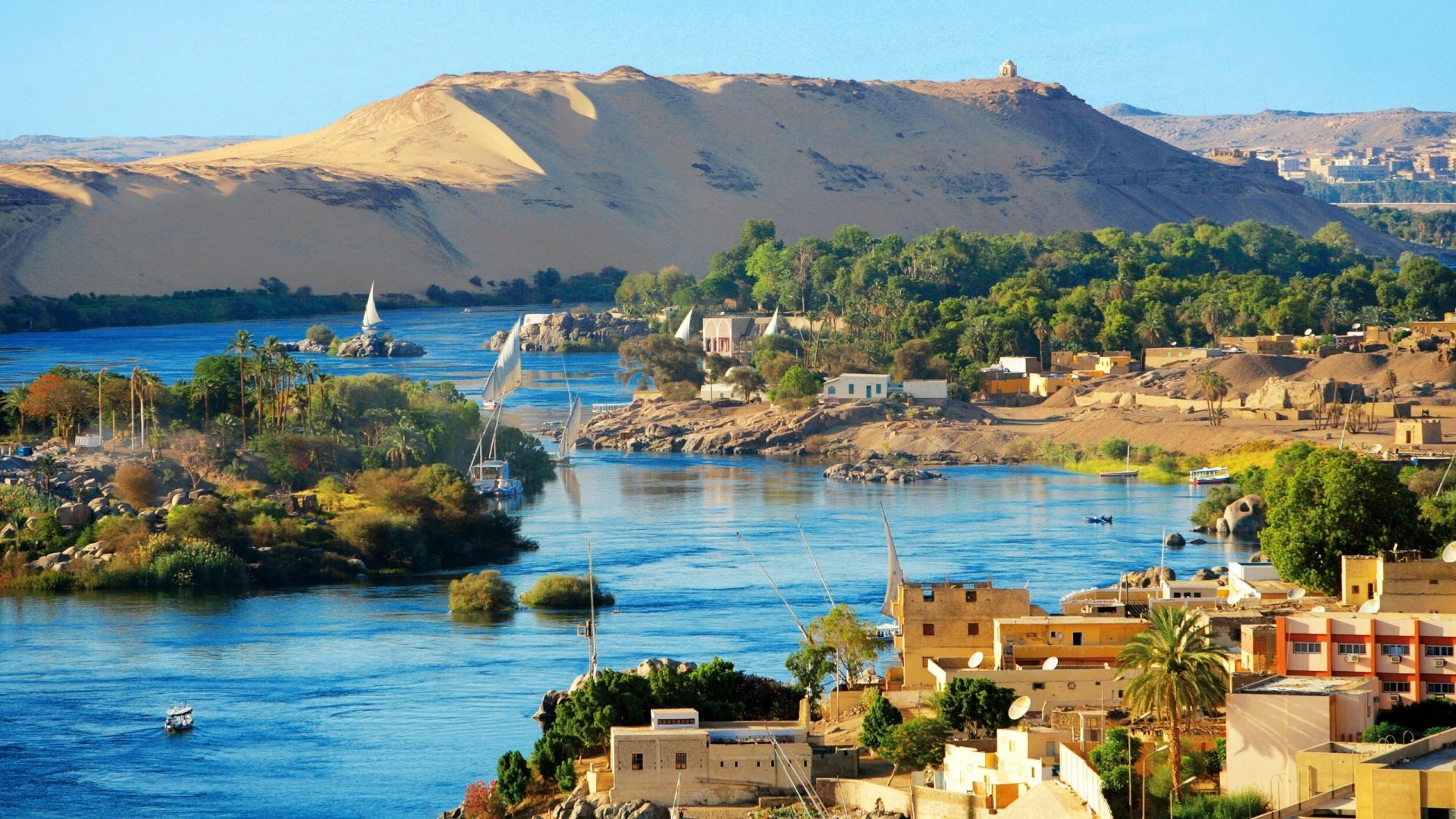 HD desktop wallpaper showcasing the man-made structures and lush greenery along the Nile River in Aswan, set against golden desert hills under a clear sky.