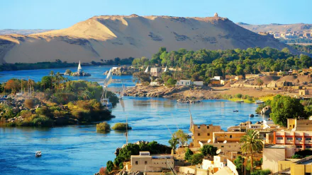 HD desktop wallpaper showcasing the man-made structures and lush greenery along the Nile River in Aswan, set against golden desert hills under a clear sky.