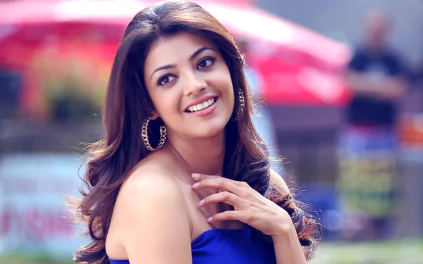 HD desktop wallpaper featuring actress and model Kajal Aggarwal, smiling with hand near her shoulder, wearing a blue outfit. The background is blurred with hints of red and green elements.