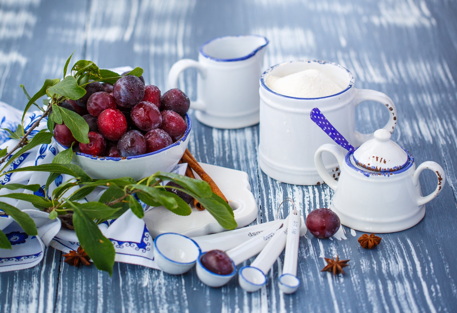 HD desktop wallpaper featuring fresh plums in a white bowl alongside white ceramic kitchenware with blue accents on a textured blue surface.