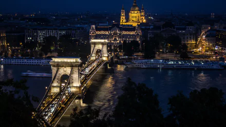 A nighttime 4K Ultra HD view of the illuminated Chain Bridge spanning a river in a cityscape, highlighting man-made architectural beauty against a dark sky.