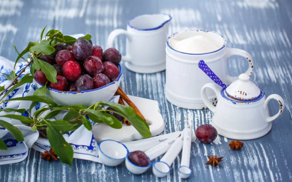 HD desktop wallpaper featuring fresh plums in a white bowl alongside white ceramic kitchenware with blue accents on a textured blue surface.
