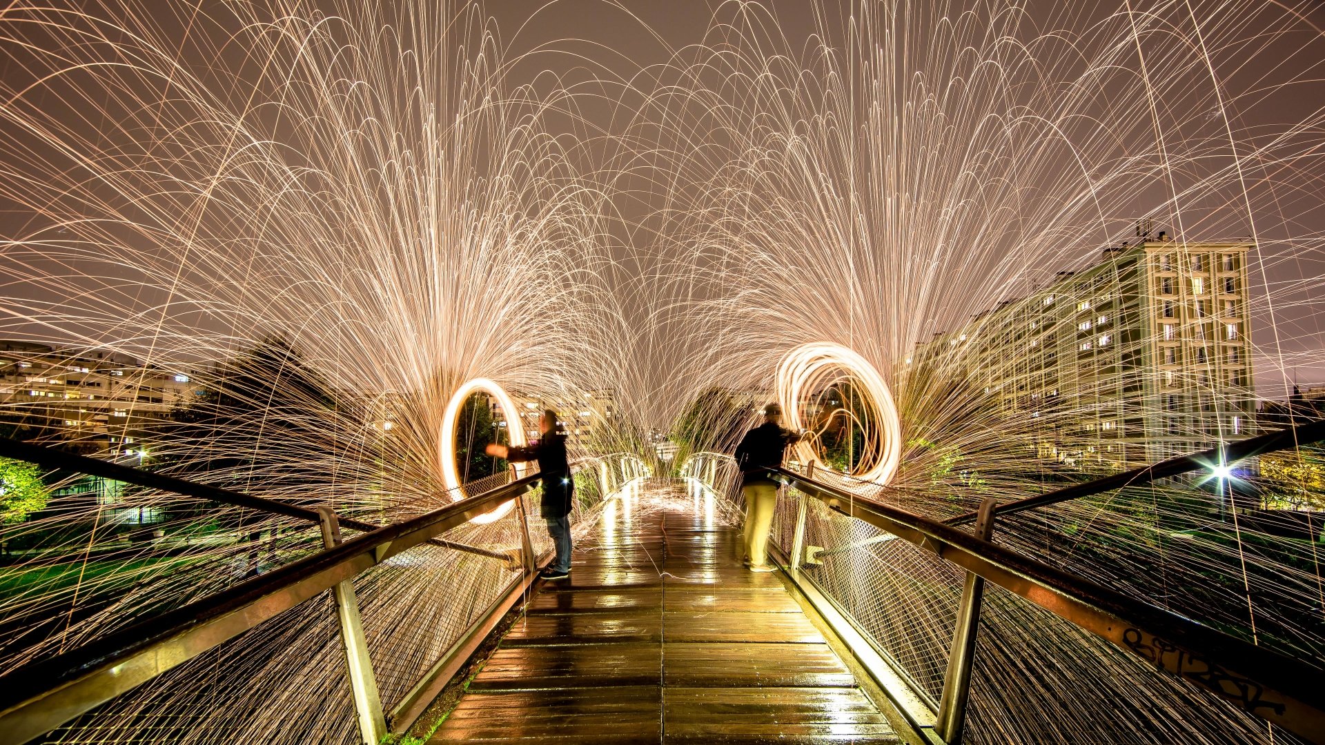 A striking image of fire juggling on a wooden bridge, showcasing vibrant sparks and dynamic light trails against a nighttime city backdrop. A stunning 4K Ultra HD desktop wallpaper.