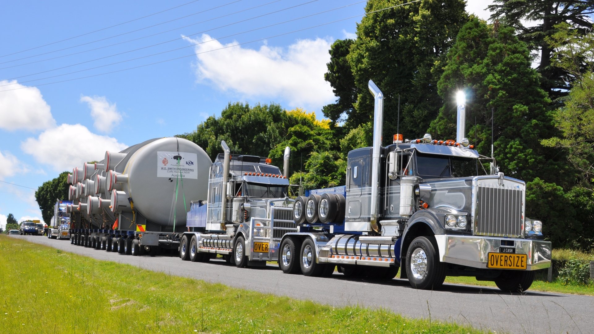 4K Ultra HD PC desktop wallpaper showing two polished Kenworth heavy-haul trucks hauling cylindrical tanks along a tree-lined country road under a bright blue sky.