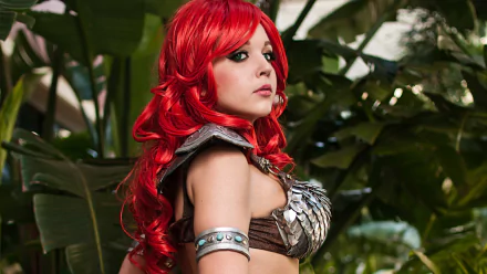 HD PC desktop wallpaper featuring a woman cosplaying as Red Sonja with vibrant red hair and armor, set against a lush green leafy background.