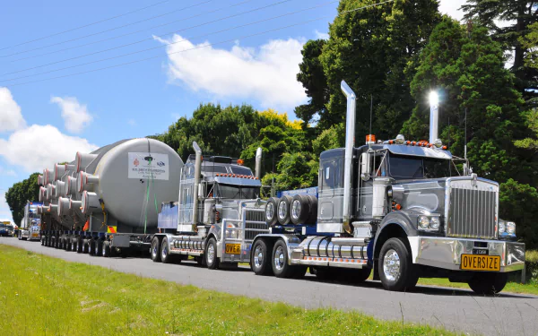 4K Ultra HD PC desktop wallpaper showing two polished Kenworth heavy-haul trucks hauling cylindrical tanks along a tree-lined country road under a bright blue sky.