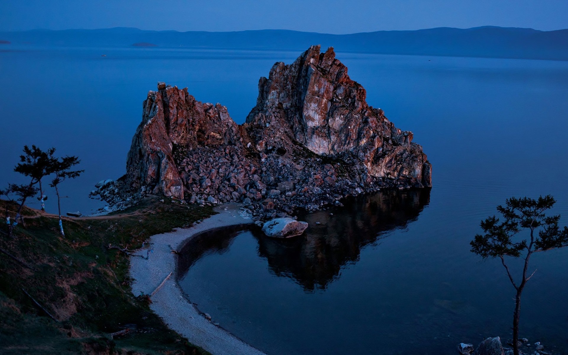 HD PC desktop wallpaper and background: nature cliff and rocky islet at twilight, crescent pebble beach and silhouetted trees reflected in calm blue water.