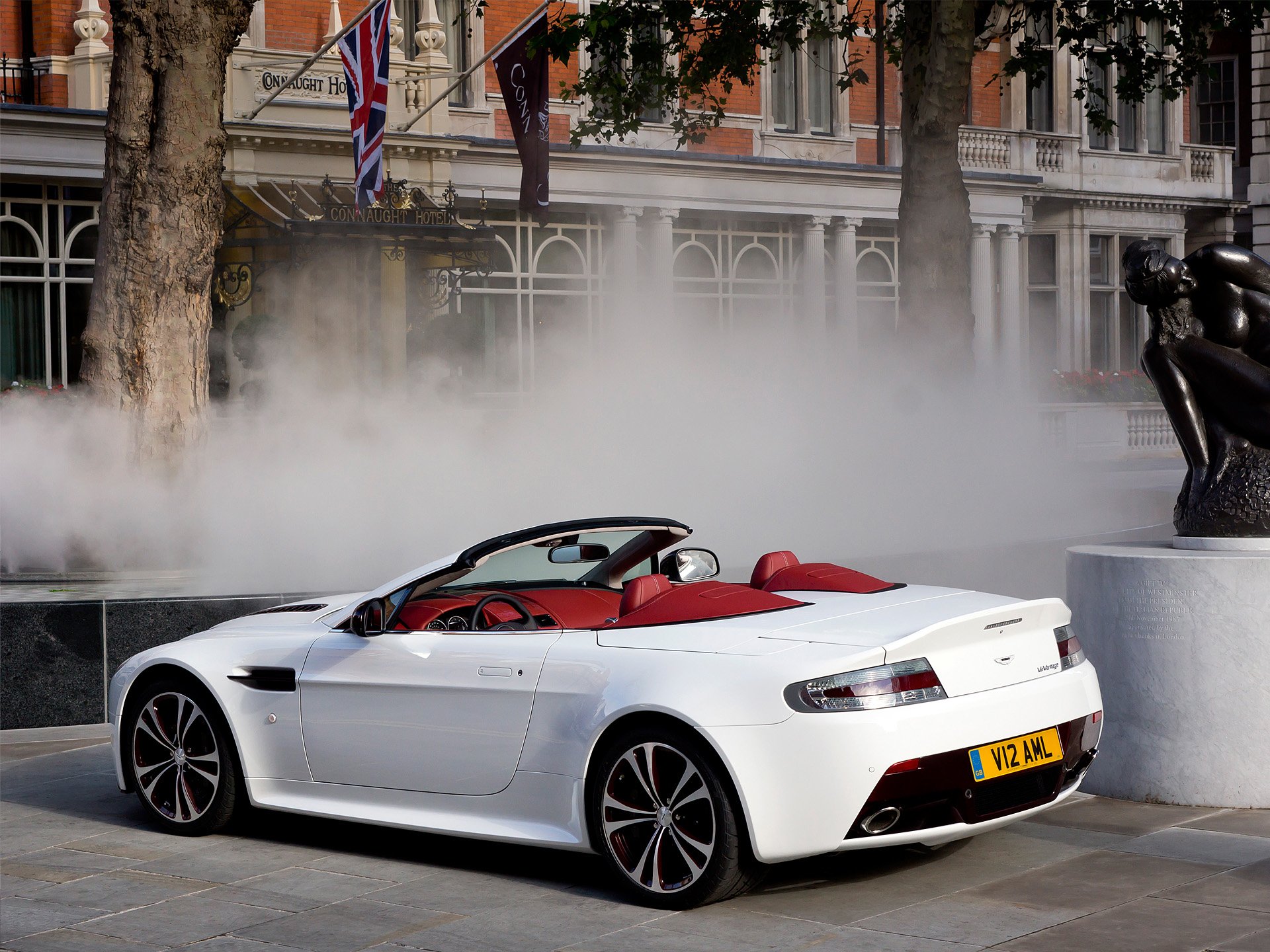 HD PC desktop wallpaper: white Aston Martin V12 Vantage convertible with red interior parked by a fountain in a city plaza.