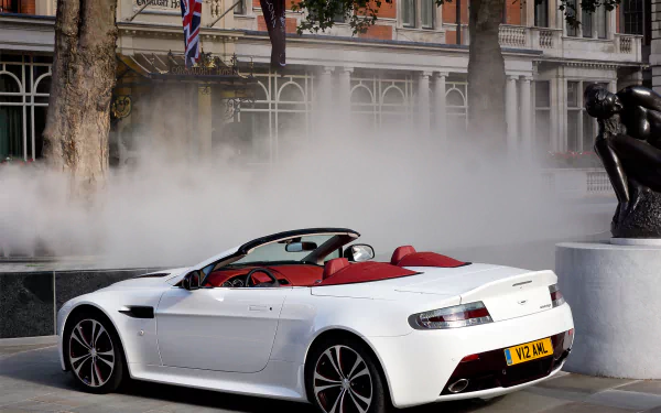 HD PC desktop wallpaper: white Aston Martin V12 Vantage convertible with red interior parked by a fountain in a city plaza.