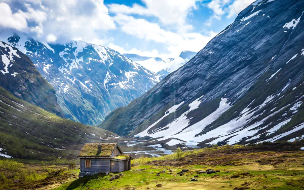 A man-made cabin nestled in a green valley surrounded by snow-capped mountains under a bright blue sky, captured in 4K Ultra HD for PC desktop background.