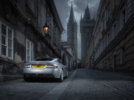 A sleek Aston Martin DBS parked on a cobblestone street, with historic buildings and a gothic tower in the background, creating a dramatic, atmospheric scene.
