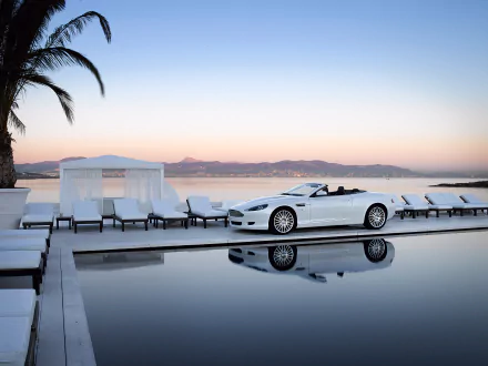 A stunning Aston Martin DB9 convertible parked by a calm pool at sunset, surrounded by lounge chairs and palm trees, creating a luxurious and serene atmosphere.