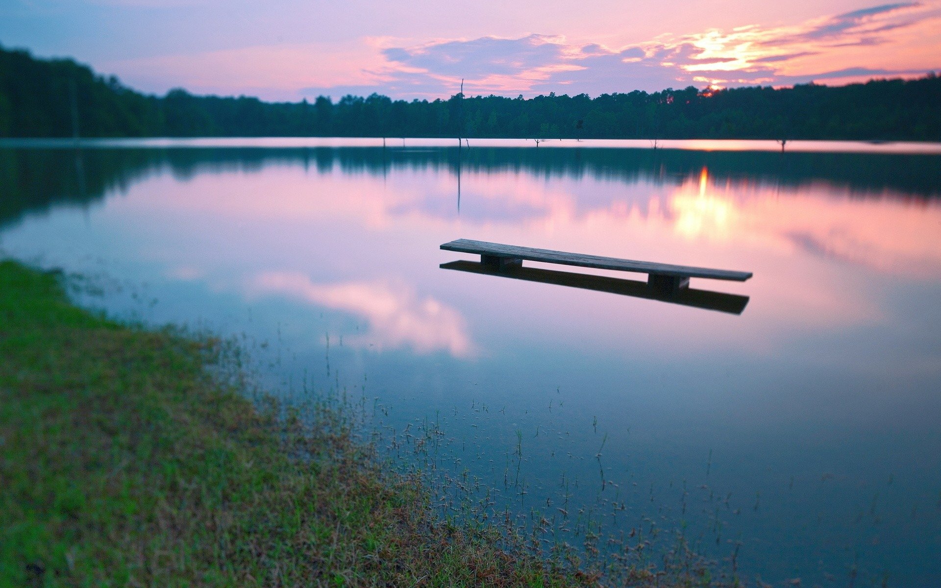 HD PC desktop wallpaper showing a tranquil lake at sunrise, with soft pink and purple skies reflecting on calm water and a floating wooden dock near the shore.