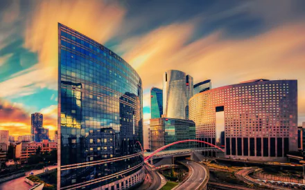 HD desktop wallpaper depicting a sunset over the modern skyscrapers and winding roads of La Défense, the business district in Paris, France.