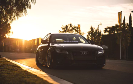 A sleek black Audi A4 parked along a sunlit road, with vibrant sunset lighting casting a warm glow, creating a striking and dynamic HD desktop wallpaper background.