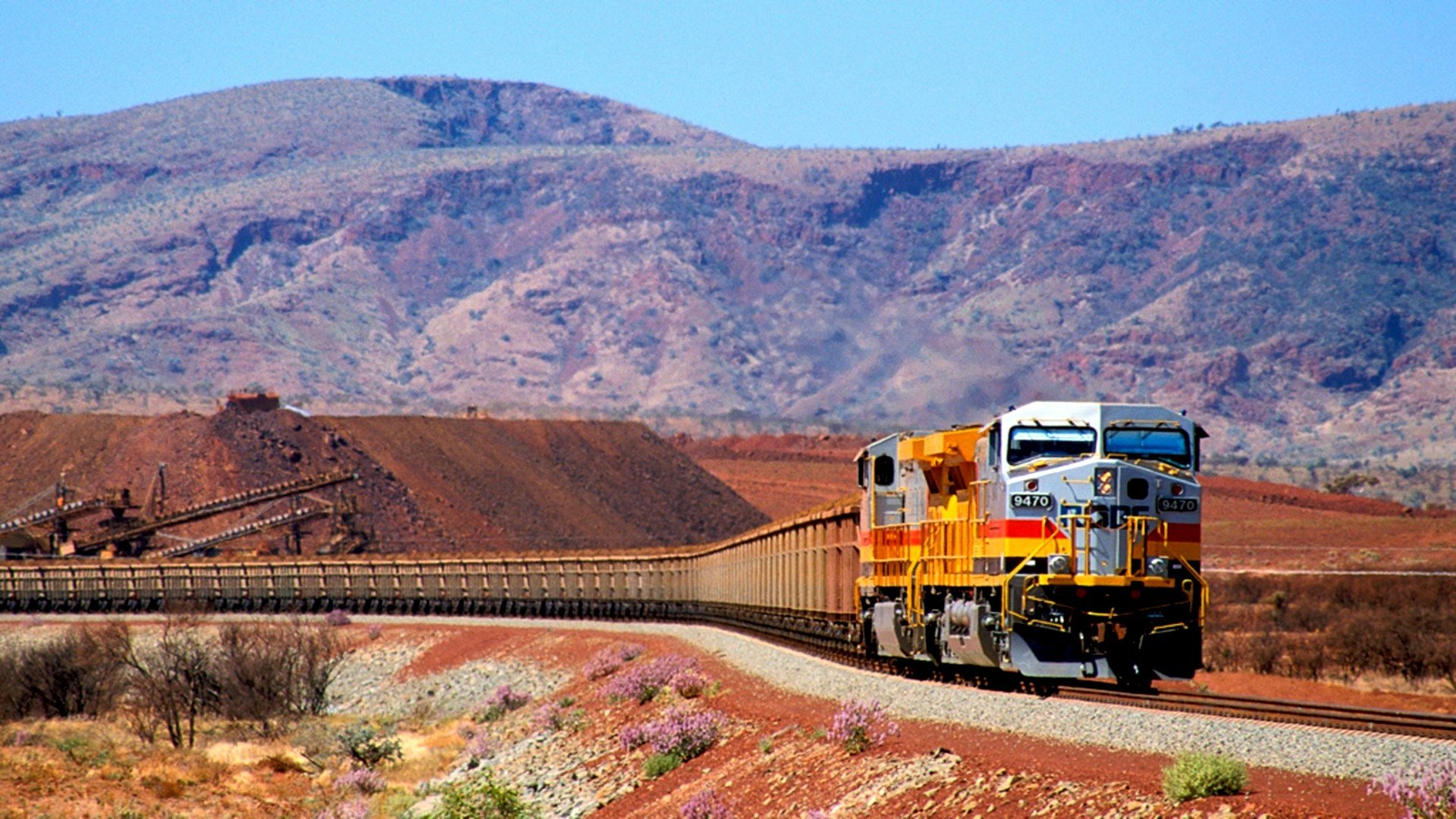 HD desktop wallpaper featuring a vibrant train traveling through a desert landscape with rugged mountains in the background under a clear blue sky.