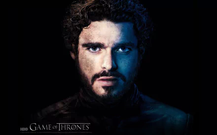 A close-up of Richard Madden as Robb Stark from the TV show Game of Thrones, featuring dramatic lighting that highlights his intense expression. Suitable as a desktop wallpaper.