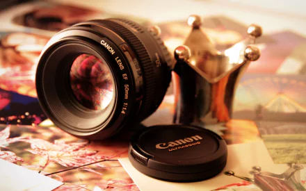 A close-up HD PC desktop wallpaper featuring a Canon camera lens, its cap, and a decorative crown-shaped object on a soft background.