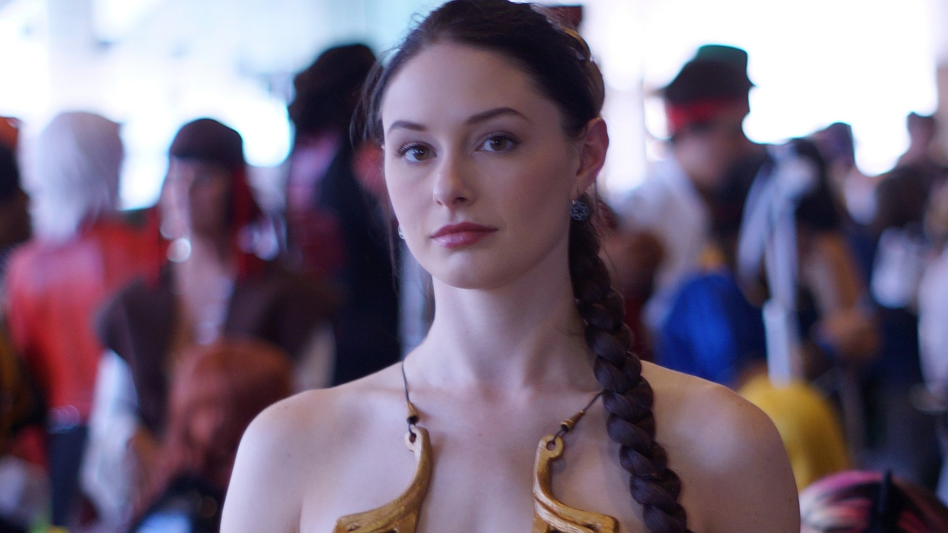 2K Quad HD PC desktop wallpaper: woman in detailed fantasy cosplay with long braid and ornate bronze costume, soft-focus convention crowd forming the background.