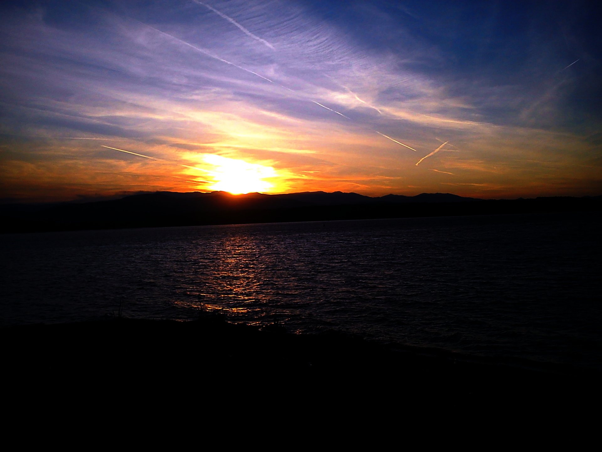 2K Quad HD PC desktop wallpaper: nature sunset — golden sun dipping behind distant hills, purple-orange sky reflected on calm water, silhouetted shoreline in the foreground.