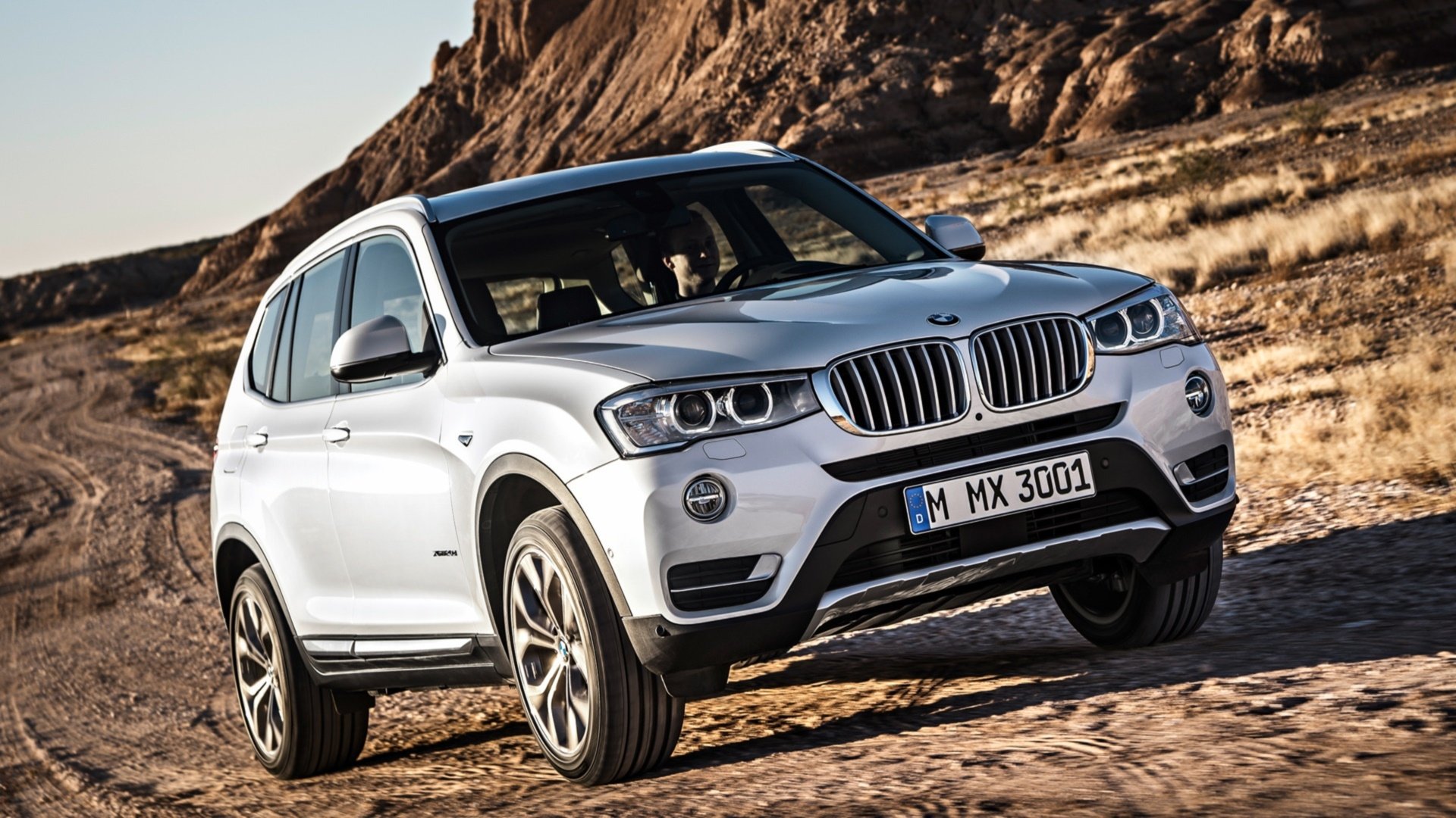 Silver 2015 BMW X3 LCI on a rocky desert road, front three-quarter view in warm light — HD PC desktop wallpaper/background of the SUV in rugged terrain.