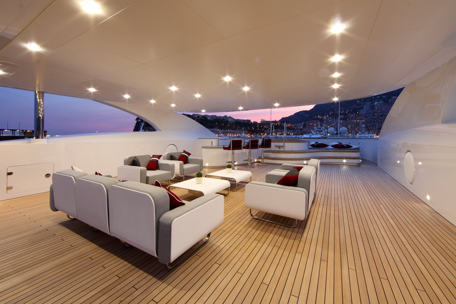 Spacious room on a luxury yacht with modern white seating, wooden deck flooring, and city lights visible at dusk, captured in 4K Ultra HD for a PC desktop wallpaper.