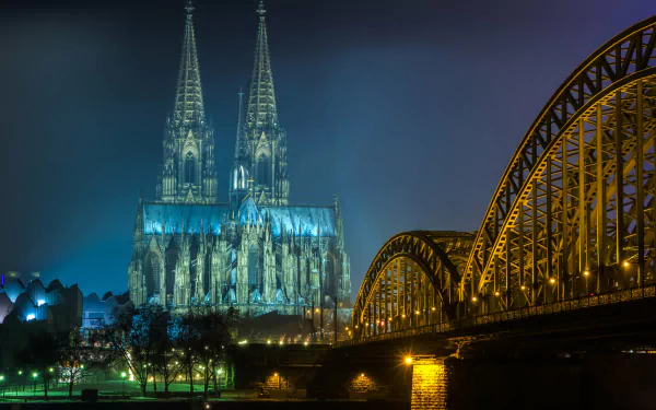 Cologne Cathedral stands majestically in the background, illuminated against a night sky, with a bridge arching gracefully in the foreground. A striking 4K Ultra HD scene.