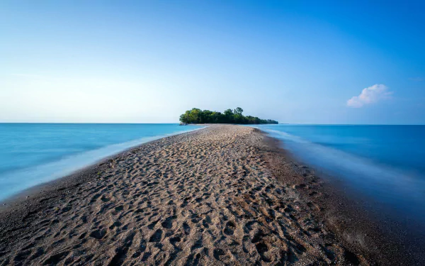 A serene landscape showcasing a sandy path leading to a lush island, surrounded by calm blue waters under a clear sky. 4K Ultra HD desktop wallpaper for nature lovers.