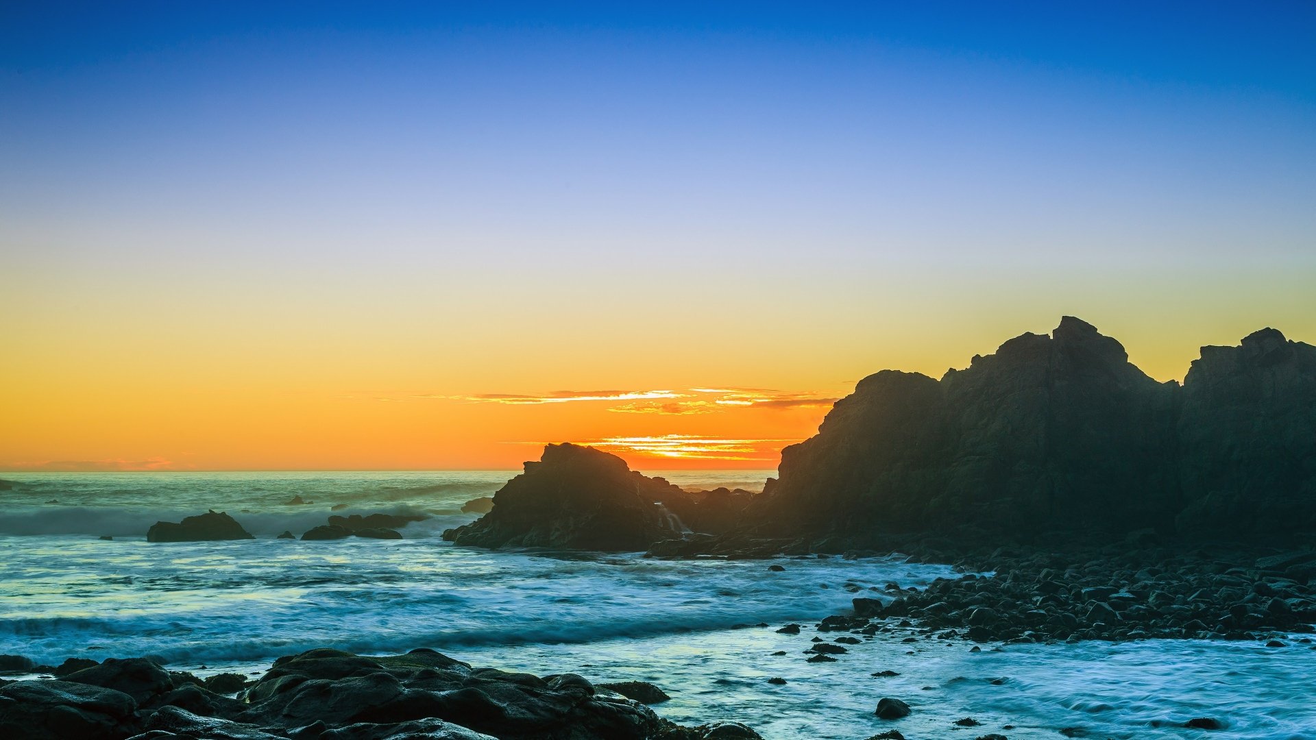 A breathtaking sunset over rocky coastline, with vibrant colors blending into the sky and waves gently crashing against the shore. This nature scene makes a stunning HD desktop wallpaper.