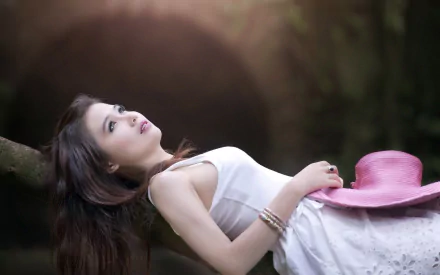 An Asian woman in a white dress with a pink hat lies on a tree branch, gazing upwards. The image is a HD desktop wallpaper and background.