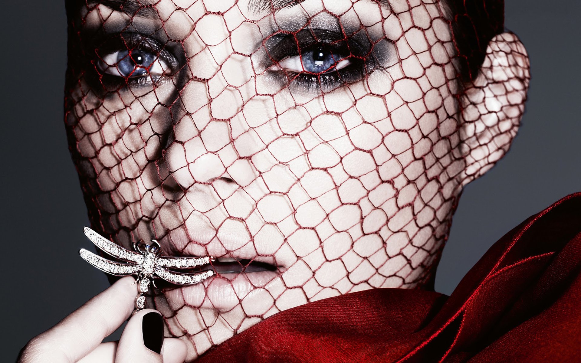Close-up HD portrait of French actress Marion Cotillard with a netted veil over her face, holding a jeweled dragonfly brooch, set against a dark background.