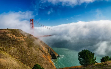 HD desktop wallpaper showcasing the man-made Golden Gate Bridge emerging from fog over water, framed by coastal cliffs and a partly cloudy blue sky.