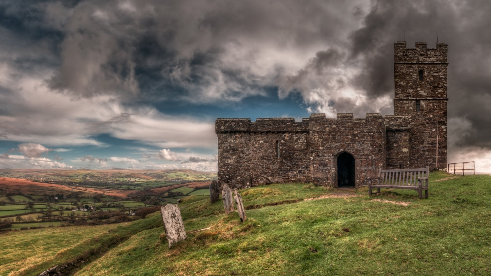 HD desktop wallpaper showing Brentor Church atop a grassy hill under dramatic, cloudy skies, embodying a striking religious and historic atmosphere.