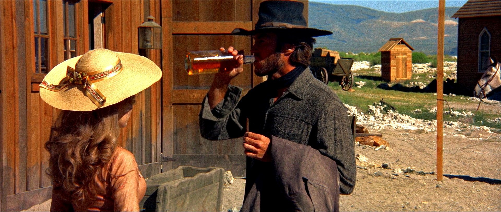 HD PC desktop wallpaper from the movie High Plains Drifter: a hat-wearing man drinks from a bottle while a woman in a sunhat stands by a wooden frontier building and distant hills.