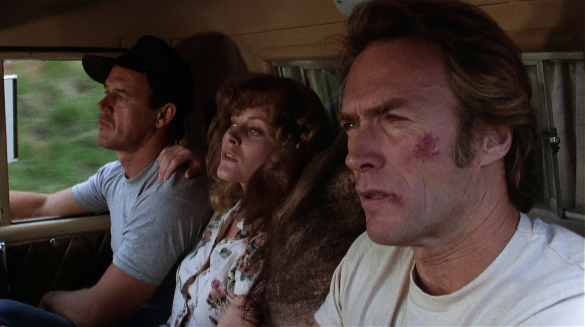 HD PC desktop wallpaper from the movie Every Which Way but Loose: three tense passengers in a pickup — driver, woman in a floral blouse, and a man with a bruise on his cheek staring ahead.