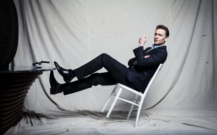 HD desktop wallpaper of a man in a black suit reclining on a white chair with legs raised, smiling against a draped studio backdrop.