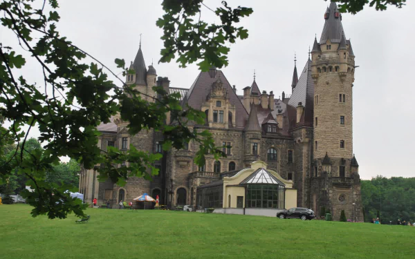 HD PC desktop wallpaper of the man-made Moszna Castle: stone turrets and ornate façade framed by leafy branches above a wide green lawn under an overcast sky.