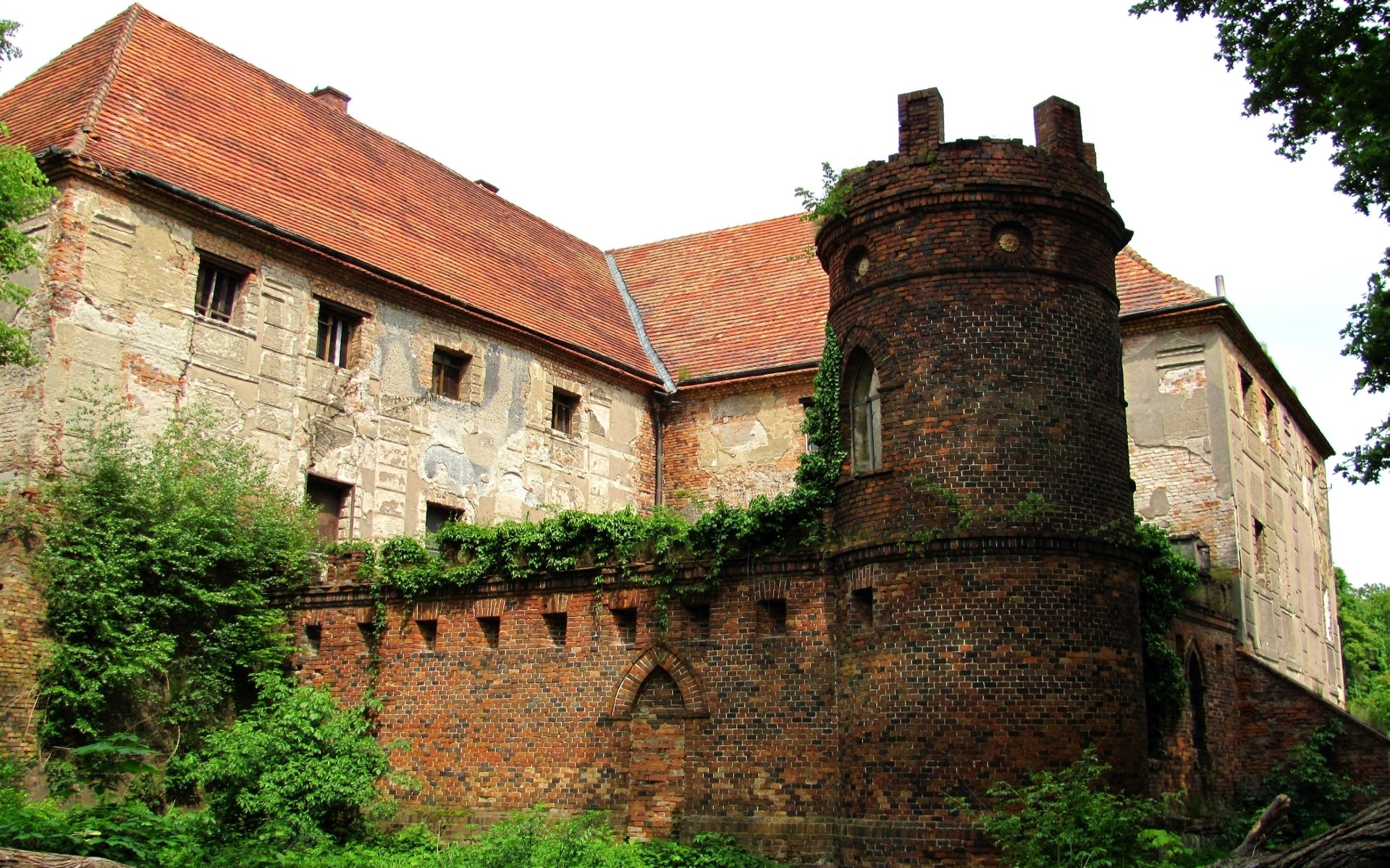 HD PC desktop wallpaper of Broniszów Castle, a man-made brick and stone fortress with a round tower, red tiled roofs and lush overgrown greenery.
