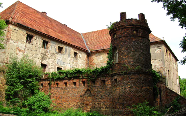 HD PC desktop wallpaper of Broniszów Castle, a man-made brick and stone fortress with a round tower, red tiled roofs and lush overgrown greenery.