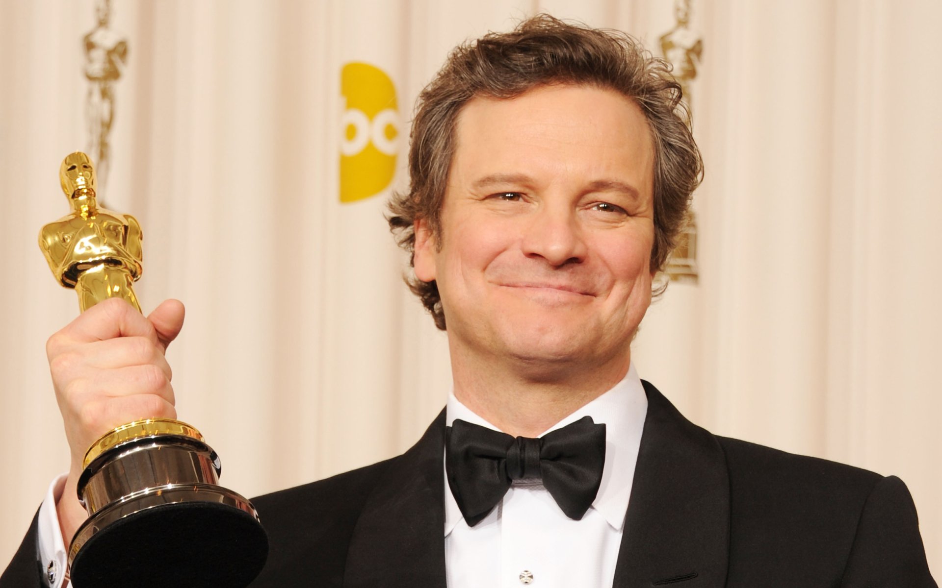 Smiling English actor in a tuxedo holding an Oscar statuette, close-up portrait styled as a 2K Quad HD PC desktop wallpaper background.