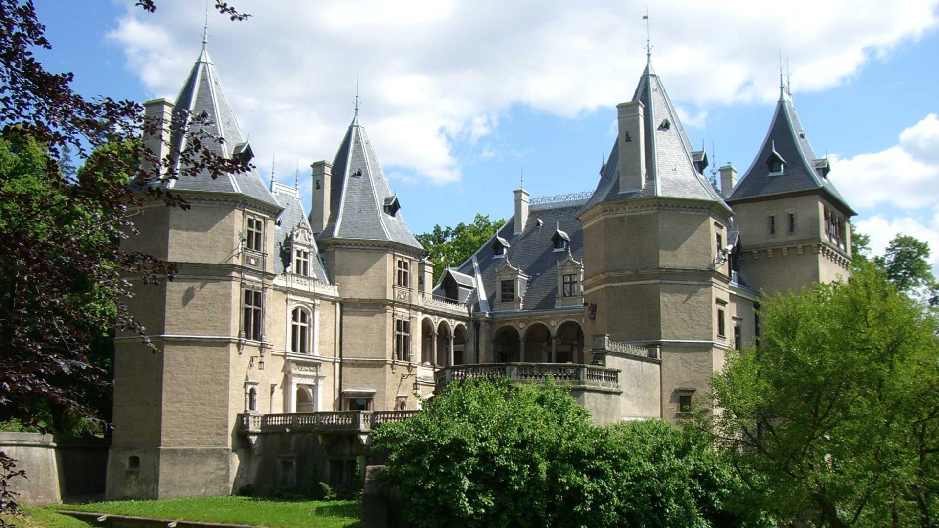 HD PC desktop wallpaper of Goluchów Castle, a man-made stone château with slate turrets and an arched gallery, set among lawns and trees under a blue sky.