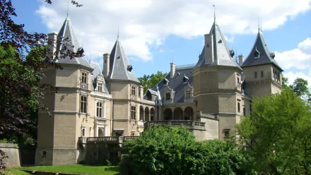 HD PC desktop wallpaper of Goluchów Castle, a man-made stone château with slate turrets and an arched gallery, set among lawns and trees under a blue sky.