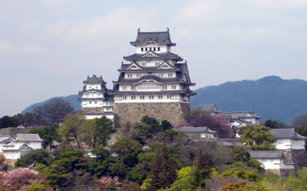 HD desktop wallpaper featuring the man-made Himeji Castle perched on a hill with surrounding greenery and distant mountains under a cloudy sky.