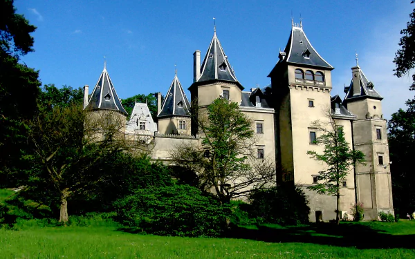Man-made Goluchów Castle with stone towers and turrets, framed by trees and a green lawn under a blue sky — HD PC desktop wallpaper/background.